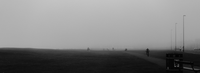 A black and white view of the links at Whitley Bay. A cyclist rides away from camera along a path, while a father and son play football in the fog and a man sits on a bench. In the background you can just make out the lower portion of the Spanish City building.