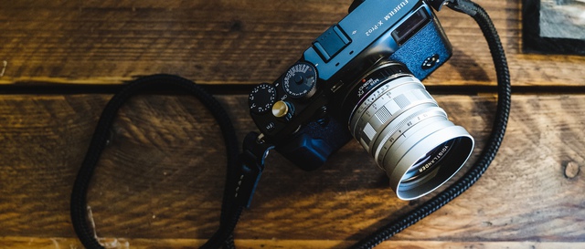 A top-down view of a black Fujifilm X-Pro2 with silver Voigtländer Nokton lens attached. The rope-/cord-style camera strap lies coiled around and beside the camera on a rustic wood table.