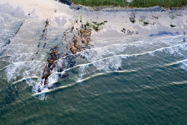 A birds-eye view looking down on Low Hauxely beach. Breaking waves create diagonal lines across the frame, while some reddish rocks poke out into the sea.