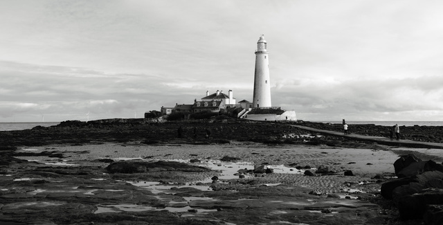 A panoramic black and white image looking across the sand and rocks to St. Mary’s Island at low tide. The island and lighthouse stand in the centre, while a couple of people walk on the causeway to the right. Wind turbines stand in the far background on the horizon, out to sea.