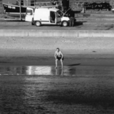 A man in swimming trunks stands at the waters edge, leaning squatting forward and watching the sea