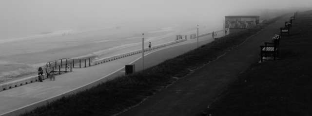 A panoramic ratio, black and white photograph, looking down from the links at Whitley Bay onto the prom and the beach. The promenade crosses diagonally bottom-left to top-right of the frame, with the grassy links in deep shadow in the foreground while the beach and sea are shrouded in fog